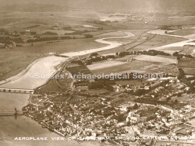 Aerial view over Shoreham-by-Sea looking north west with River Adur in foreground and South Downs with Chanctonbury Ring in distant background. Footbridge visible in foreground, railway bridge on left and old toll bridge in middleground with Lancing College and Chapel to its left. Catholic church in foreground centre left, St Mary de Haura centre right with Shoreham Grammar school in Pond Road behind the tower of St Mary's. River Adur winding through fields into distance.